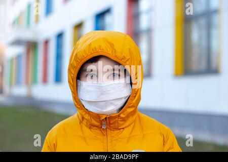 Ritratto di un ragazzo in una giacca gialla e cappuccio in una maschera medica bianca sulla strada vicino all'edificio con finestre multicolore.concetto di malattia quara Foto Stock