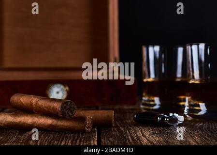 Ancora vita con tre sigari cubani, due bicchieri di whisky o rum, più leggero e scatola di legno con igrometro. Vecchio tavolo in legno e sfondo nero. Foto Stock