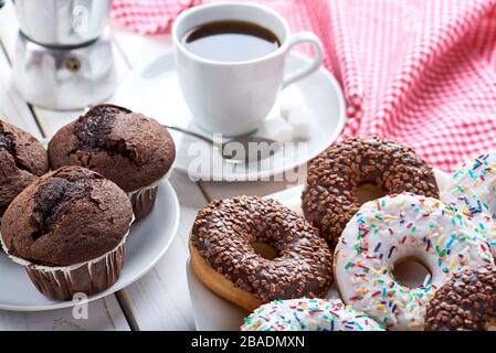 Colazione dolce con muffin al cioccolato e ciambelle, una tazza di caffè nero su tavola di legno bianco. Strofinaccio rosso e spazio per il testo. Foto Stock