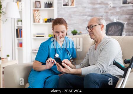 Uomo anziano in casa di cura con stampelle che mostrano qualcosa sul telefono mobile al suo ospite di salute. Foto Stock