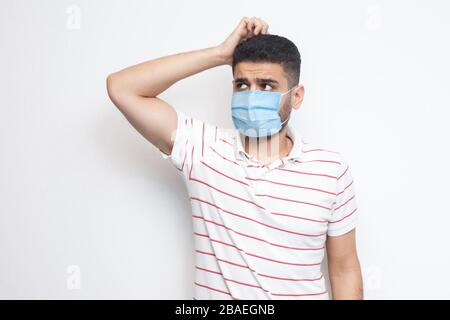 Ritratto di query giovane uomo con maschera medica chirurgica in una t-shirt a righe in piedi graffiare la testa, planando e pensando a qualcosa. Interni s Foto Stock