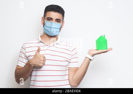 Ti sentirai come a casa, in tutta sicurezza. L'uomo positivo con maschera medica chirurgica che tiene la piccola casa di carta verde, propone di essere a casa sul tempo di quarantena, pollici in su. Foto Stock