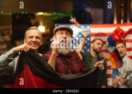 Uomini maturi con bandiera tedesca che si rallegrano per la loro squadra di calcio mentre guardano la partita nel bar sportivo con gli appassionati americani in background Foto Stock