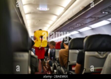 Foto sfocata dell'interno di una cabina di aeroplano con la stewardess che fornisce le istruzioni in caso di emergenza. Giubbotto di emergenza giallo. Foto Stock