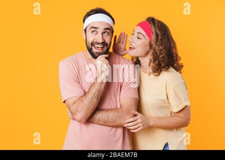 Ritratto di giovane coppia atletica che indossa le fasce della testa che si domandano mentre sussurrano segreto o pettegolezzo isolato su sfondo giallo Foto Stock