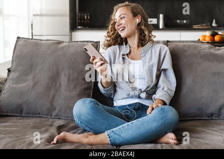 Allegra giovane donna bionda con i capelli che ascolta musica con le cuffie mentre siede su un divano a casa e tiene il telefono cellulare Foto Stock