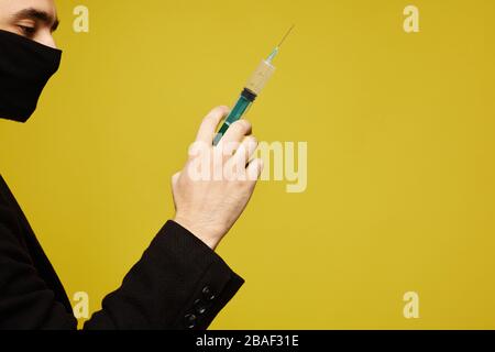 Vista laterale del bell'uomo in maschera protettiva nera che tiene una siringa in mano, isolato sullo sfondo giallo con spazio di copia per il testo. Foto Stock