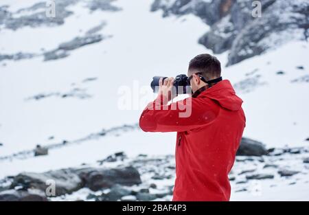 Vista posteriore di un uomo che scatta foto con una fotocamera professionale. Ragazzo che indossa una giacca rossa mentre fotografa la natura invernale. Concetto di viaggio, trekking e fotografia professionale. Foto Stock