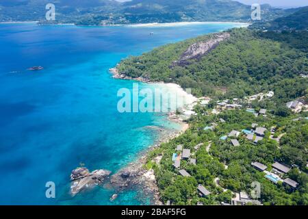 Vista panoramica aerea della baia di Anse Petite con villaggio, colline boscose e mare turchese, Mahe, Seychelles Foto Stock