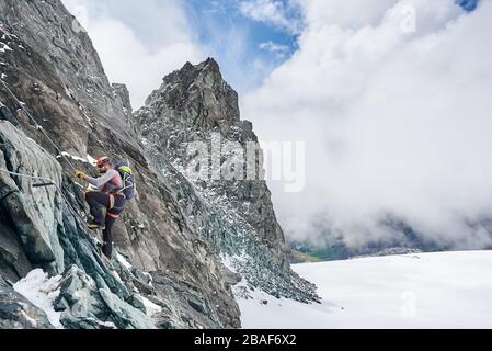 Vista laterale dell'arrampicatore in casco di sicurezza con corda fissa mentre sale montagna rocciosa con cielo bellissimo sullo sfondo. Alpinista con formazione naturale di roccia di arrampicata dello zaino. Concetto di alpinismo Foto Stock