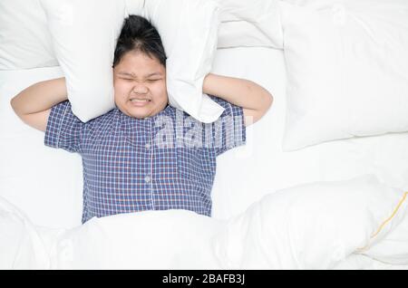 Ragazzo grasso obeso sdraiato sul letto coprendo la testa con cuscino perché troppo forte fastidioso rumore. Il bambino irritato che soffre dai vicini rumorosi, provando a dormire Foto Stock