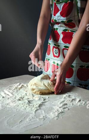 Le mani della donna impastano l'impasto e lo stampano con il Flour bianco sul tavolo da cucina su sfondo scuro. Beam Light Falls sul tavolo al buio. Foto Stock