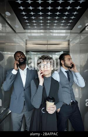 Gruppo di tre giovani manager interculturali con smartphone che chiamano i clienti all'interno dell'ascensore durante la pausa caffè Foto Stock