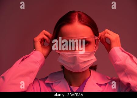 Giovane virologo o farmacista in occhiali protettivi e whitecoat mettere maschera sul viso prima di iniziare a lavorare Foto Stock