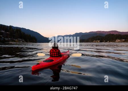 Avventurosa ragazza che pagaia su un luminoso kayak rosso in acque calme dell'oceano durante un tramonto vibrante e colorato. Preso in Indian Arm, Deep Cove, North Vancou Foto Stock