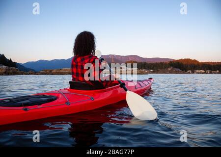 Avventurosa ragazza che pagaia su un luminoso kayak rosso in acque calme dell'oceano durante un tramonto vibrante e colorato. Preso in Indian Arm, Deep Cove, North Vancou Foto Stock