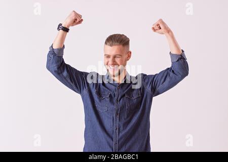 Studio ha girato l'uomo giovane hippster che celebra la vittoria mentre si levano in piedi sullo sfondo bianco. Bell'uomo felice in camicia denim che tiene le braccia sollevate e mq Foto Stock