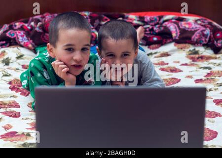 due piccoli fratelli in pigiama stanno guardando un cartone animato su un computer portatile. Bambini divertenti che si divertano con cartoni animati su notebook, laptop o computer. Foto Stock