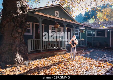 Una donna con un bambino sta stando in piedi vicino ad una casa su foglie gialle Foto Stock