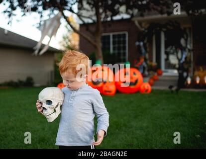 Toddler che tiene testa scheletro di fronte al decor di Halloween esterno Foto Stock