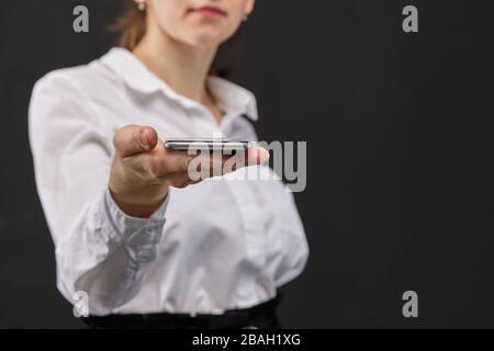 Ragazza in una camicia bianca tiene fuori la sua mano con uno smartphone su uno sfondo nero. Primo piano. Foto Stock