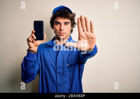 Giovane meccanico che indossa uno smartphone con tenuta uniforme su sfondo bianco isolato con mano aperta e segno di stop con un espresso serio e sicuro Foto Stock