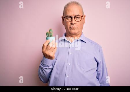 Medio età bel ragazzo tenente pentola piccola pianta cactus su sfondo rosa isolato con un'espressione sicura sul viso intelligente pensiero serio Foto Stock