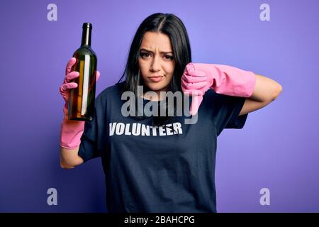 Giovane donna brunetta che indossa volontario t-shirt raccolta bottiglia di vetro su sfondo viola con faccia arrabbiata, segno negativo che mostra dislike con i pollici Foto Stock