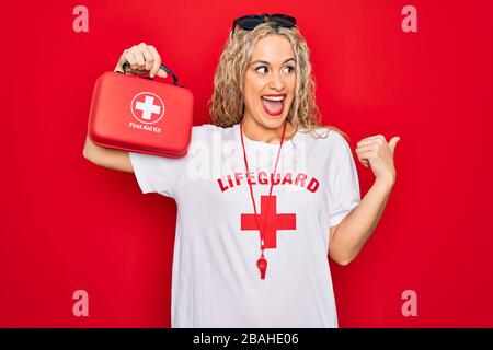 Bella donna bagnino bionda con t-shirt con croce rossa e fischio tenere kit di pronto soccorso puntando il pollice fino al lato sorridente felice con l'ope Foto Stock