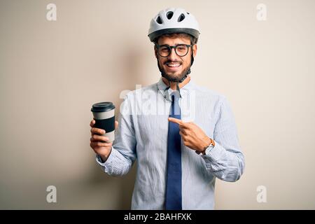 Giovane uomo d'affari che indossa occhiali e casco da bici bere tazza di caffè molto felice puntando con mano e dito Foto Stock