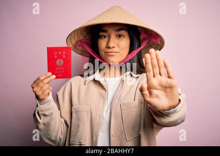 Giovane donna tailandese che indossa tradizionale cappello conico asiatico in possesso del giappone passaporto giapponese con mano aperta facendo stop segno con serio e fiducioso expre Foto Stock