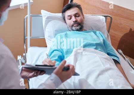 Paziente maturo a bearded nella garzatura dell'ospedale che giace nel letto e che firma l'accordo medico mentre dà il consenso all'operazione chirurgica Foto Stock