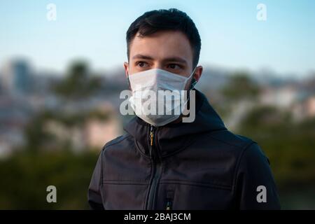 Giovane uomo isolato che indossa la maschera per proteggersi dal virus corona guarda verso con gli edifici sfocati in background Foto Stock