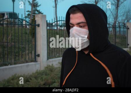 Giovane uomo isolato che indossa la maschera per la protezione e una felpa con cappuccio che riposa in un parco Foto Stock