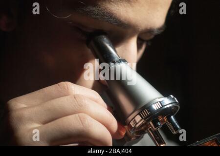 Giovane studente di scienza che fa ricerca guardando attraverso il suo microscopio a tarda notte a casa sua Foto Stock