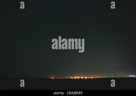 Notte Starry Sky sopra il campo e le luci gialle della città sullo sfondo. Stelle luminose naturali sopra il suolo scuro. Foto Stock