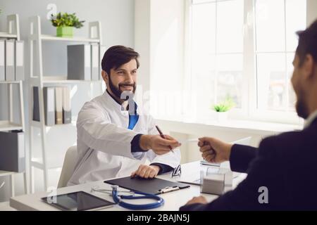 Il medico sicuro dà al paziente una prescrizione mentre si siede ad un tavolo in un ufficio della clinica. Foto Stock