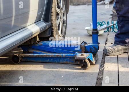 Sollevamento di auto civili argentate con vecchio martinetto idraulico a pavimento in un'officina di auto all'aperto. Procedura di sostituzione degli pneumatici per stagione. Primo piano con messa a fuoco selettiva e ba Foto Stock