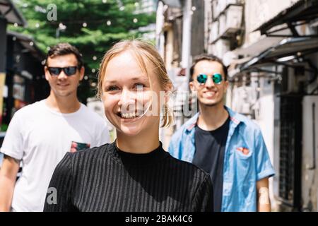 Bionda ragazza sorridente di fronte a due amici su una strada con alberi e luci appesi in una città Foto Stock