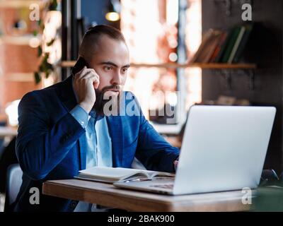 Un uomo d'affari con bearded serio conduce le conversazioni telefoniche su un telefono mobile al calcolatore. L'uomo infelice si lamenta al telefono. Emitations di rabbia in Foto Stock