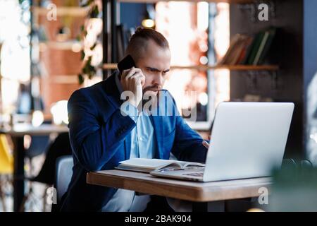 Un uomo d'affari con bearded serio conduce le conversazioni telefoniche su un telefono mobile al calcolatore. L'uomo infelice si lamenta al telefono. Emitations di rabbia in Foto Stock