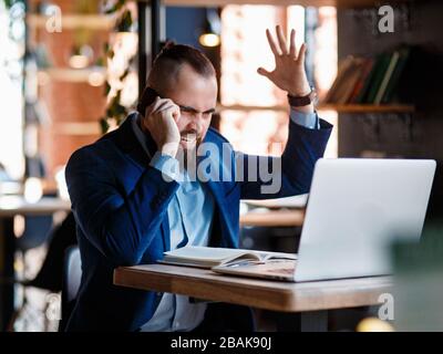 Un uomo d'affari con bearded serio conduce le conversazioni telefoniche su un telefono mobile al calcolatore. L'uomo infelice si lamenta al telefono. Emitations di rabbia in Foto Stock