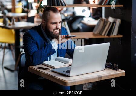 Un uomo d'affari con bearded serio conduce le conversazioni telefoniche su un telefono mobile al calcolatore. L'uomo infelice si lamenta al telefono. Emitations di rabbia in Foto Stock