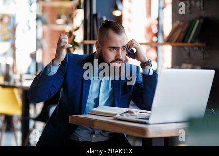 Un uomo d'affari con bearded serio conduce le conversazioni telefoniche su un telefono mobile al calcolatore. L'uomo infelice si lamenta al telefono. Emitations di rabbia in Foto Stock