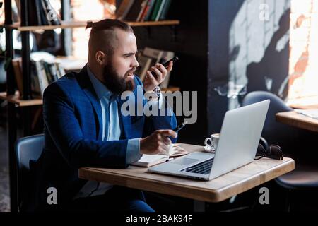 Un uomo d'affari con bearded serio conduce le conversazioni telefoniche su un telefono mobile al calcolatore. L'uomo infelice si lamenta al telefono. Emitations di rabbia in Foto Stock