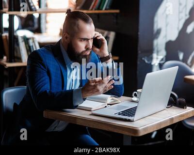 Un uomo d'affari con bearded serio conduce le conversazioni telefoniche su un telefono mobile al calcolatore. L'uomo infelice si lamenta al telefono. Emitations di rabbia in Foto Stock