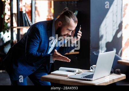 Un uomo d'affari con bearded serio conduce le conversazioni telefoniche su un telefono mobile al calcolatore. L'uomo infelice si lamenta al telefono. Emitations di rabbia in Foto Stock