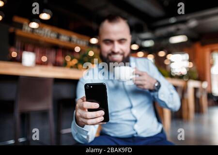 Al mattino, l'imprenditore segue le notizie, il prezzo di borsa via telefono e bevande caffè. Giovane investitore. L'uomo barbuto con un sorriso guarda dentro Foto Stock