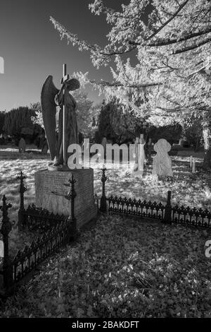 Una statua di un angelo che tiene una croce si affaccia su più tombe in un cimitero. Presa in infrarosso vicino (720 nm). Foto Stock