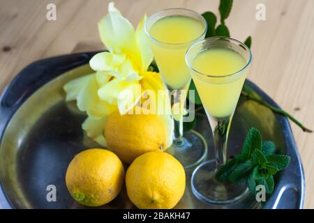 Limoncello in eleganti bicchieri di liquore con limoni, menta e rosa gialla su piastra di acciaio su tavola di legno. Liquore siciliano Foto Stock